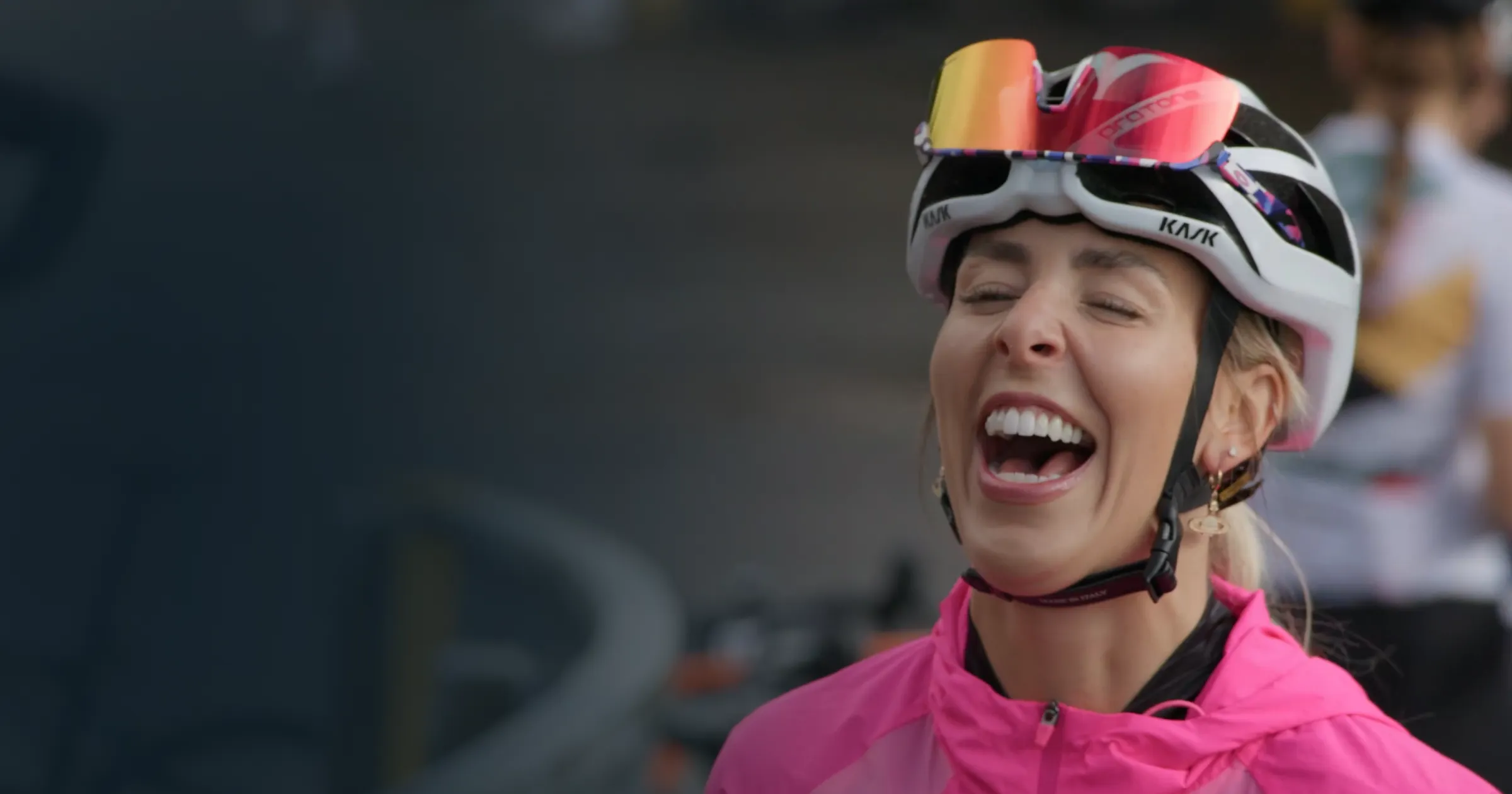 A cyclist in a pink jacket and helmet laughs joyfully. 
